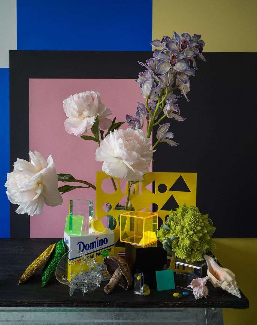 Still life photograph featuring white peonies and purple orchids against a colorful geometric background, with a sugar packet box and crystals.