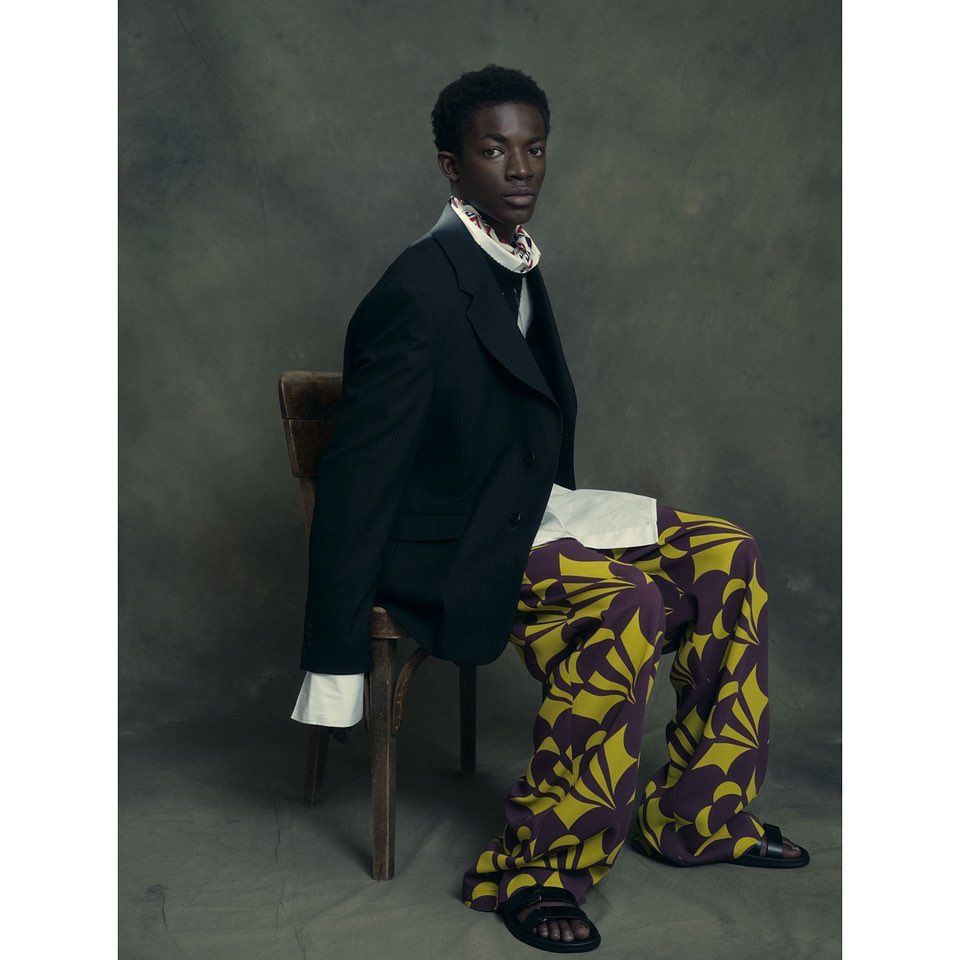 Man sits in a studio on a wooden chair wearing a black suit jacket, printed trousers, and sandals against a grey backdrop.
