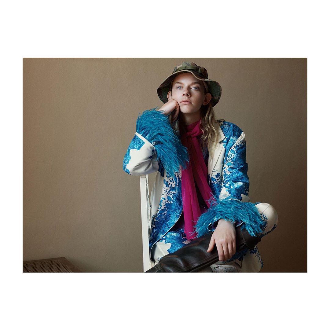 Adult female model posing in a studio. She is wearing a patterned jacket with feather cuffs, a scarf, a bucket hat, and black boots.