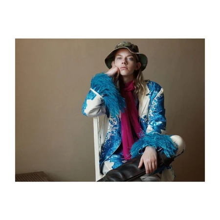 Adult female model posing in a studio. She is wearing a patterned jacket with feather cuffs, a scarf, a bucket hat, and black boots.