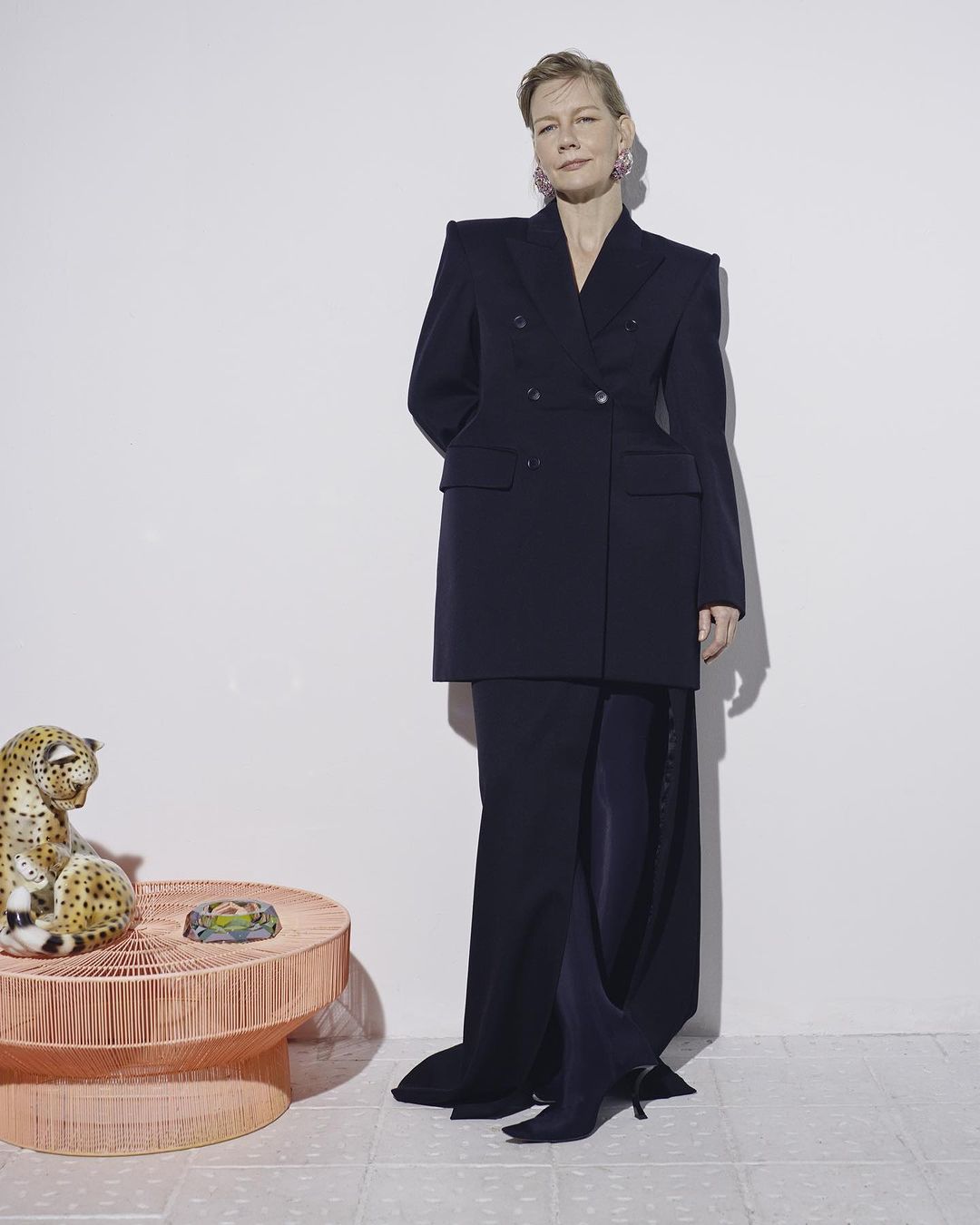 Stylish woman in oversized navy suit jacket and long skirt, standing next to a ceramic leopard statue in a minimalist studio setting.