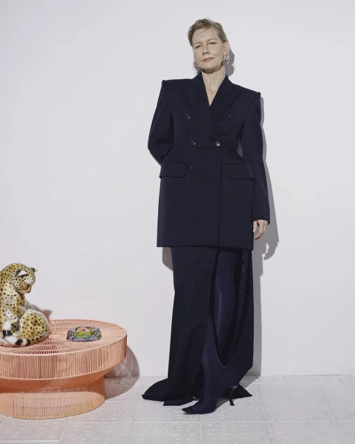 Stylish woman in oversized navy suit jacket and long skirt, standing next to a ceramic leopard statue in a minimalist studio setting.