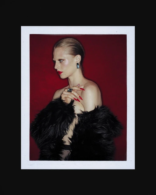 Profile view of a blonde female model with long red nails wearing a black fur coat, red lipstick, and statement earrings against a red backdrop.