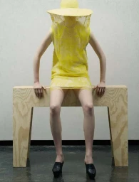 Model wearing yellow dress and veiled hat sitting on wooden bench by photographer Ralph Mecke