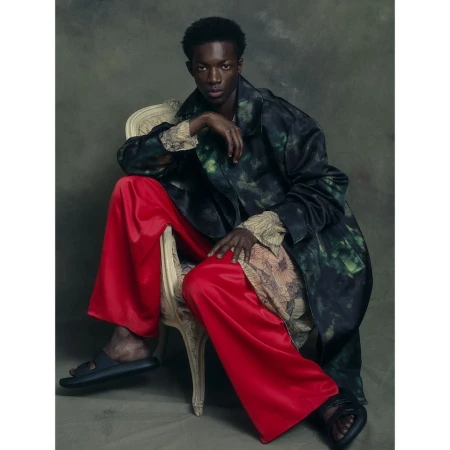Young Black man seated on an ornate chair wearing bright red trousers and a dark patterned coat by Ralph Mecke