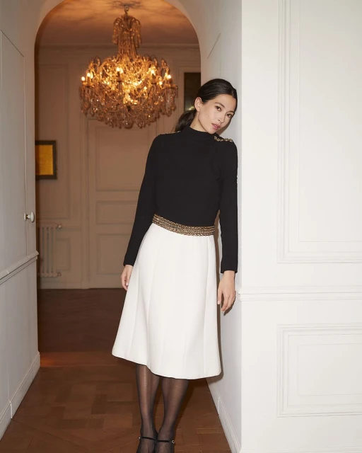Elegant Asian woman in a black top and cream skirt stands in a hallway with a chandelier, posing for a fashion shot in an indoor setting.