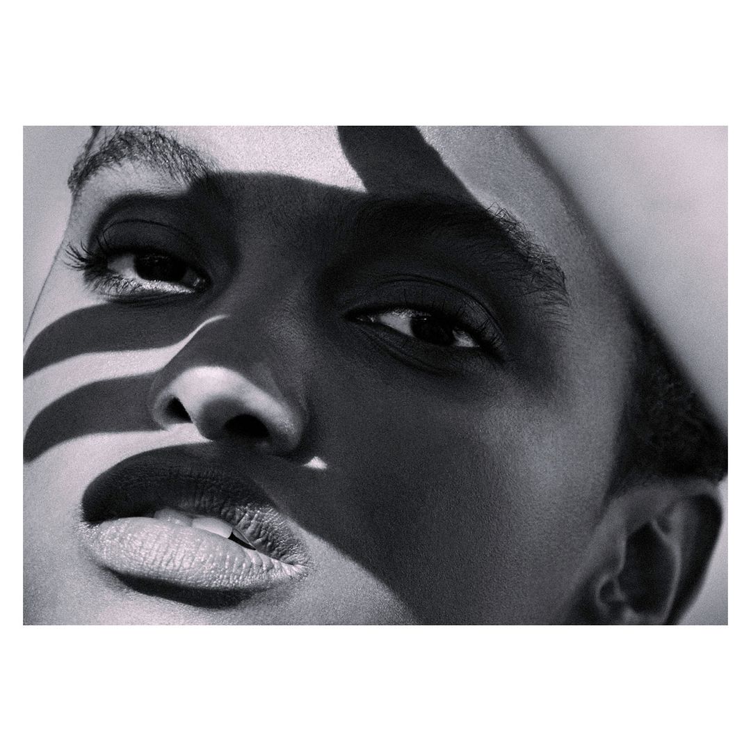 Close-up portrait of a Black woman's face with shadows creating zebra-like stripes across her forehead and left cheek in black and white.