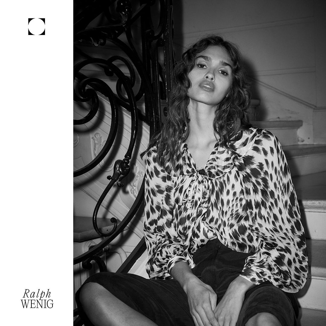 Black and white fashion portrait of a young woman with long dark hair, wearing an animal print blouse, sitting on stairs indoors.