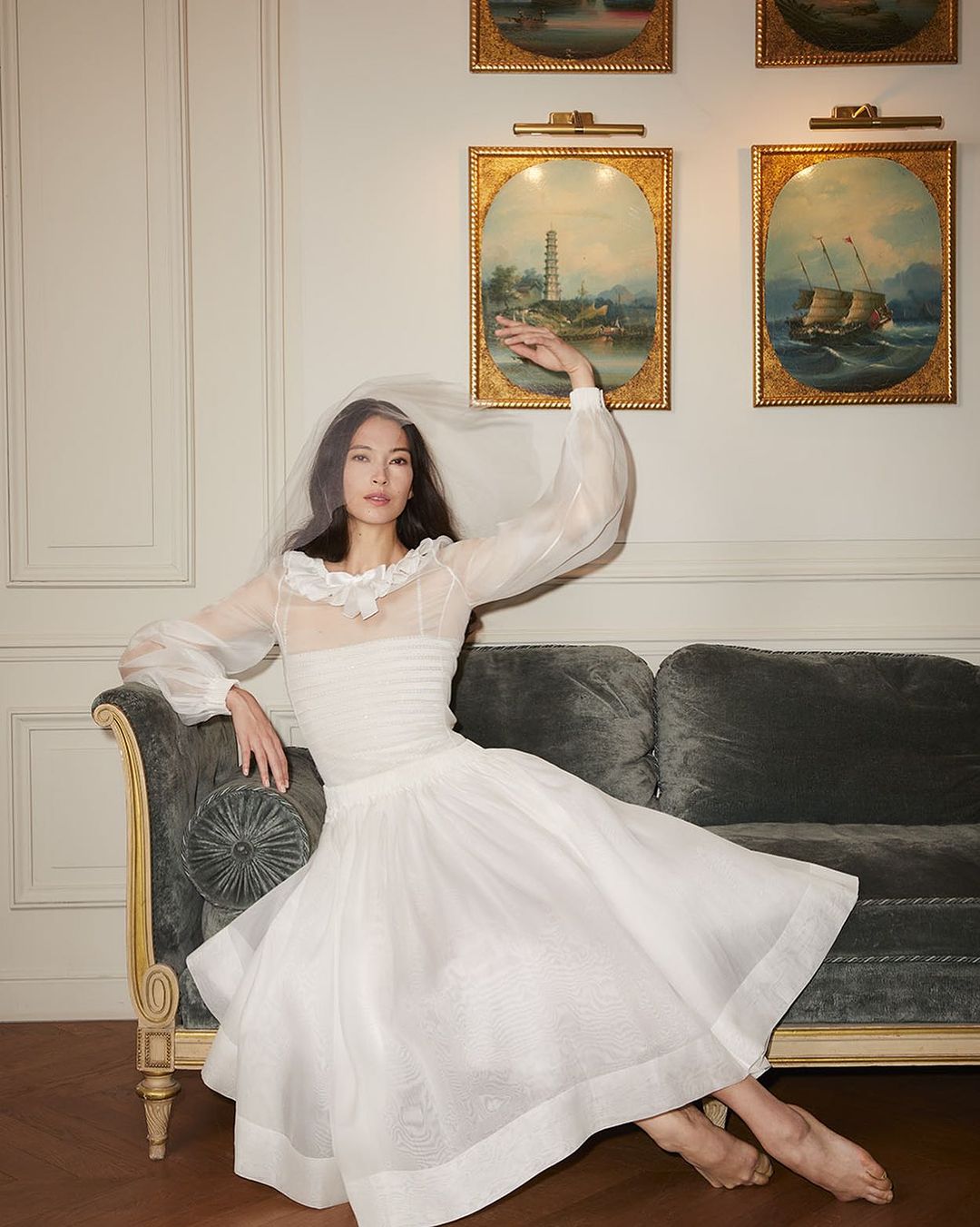 Young Asian woman in white dress and veil sits on a sofa in a bright indoor setting. She has one arm raised to the side.