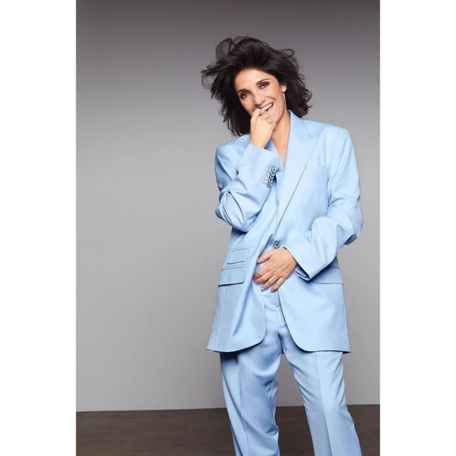 Brunette woman smiles in a light blue suit against a gray background in a medium studio shot showcasing a modern fashion portrait.