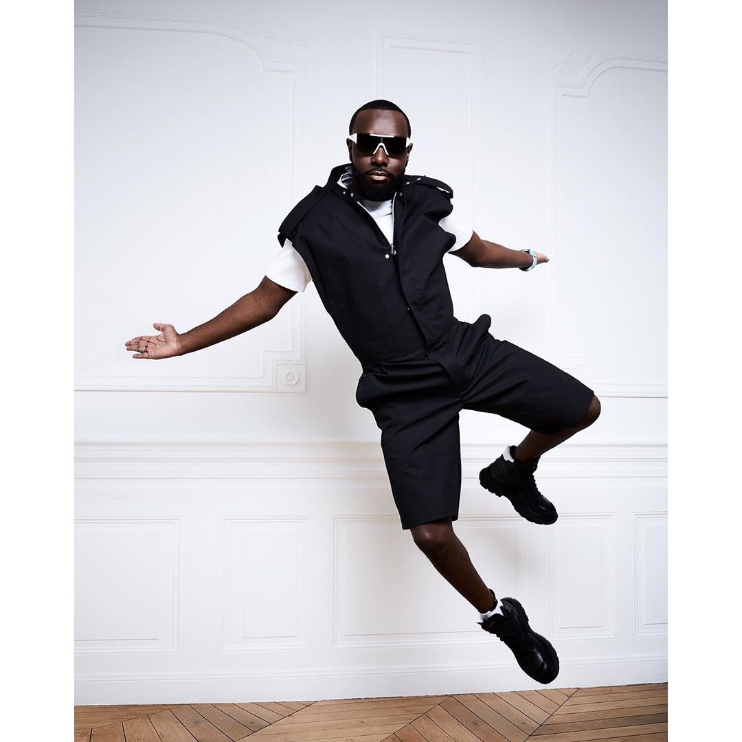An African-American man in black shorts and vest, white T-shirt, and black sunglasses leaps in the air in a photo studio.