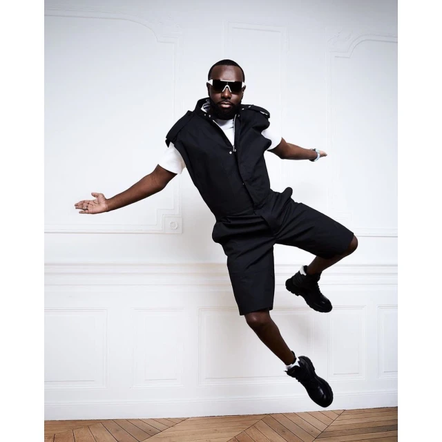 An African-American man in black shorts and vest, white T-shirt, and black sunglasses leaps in the air in a photo studio.