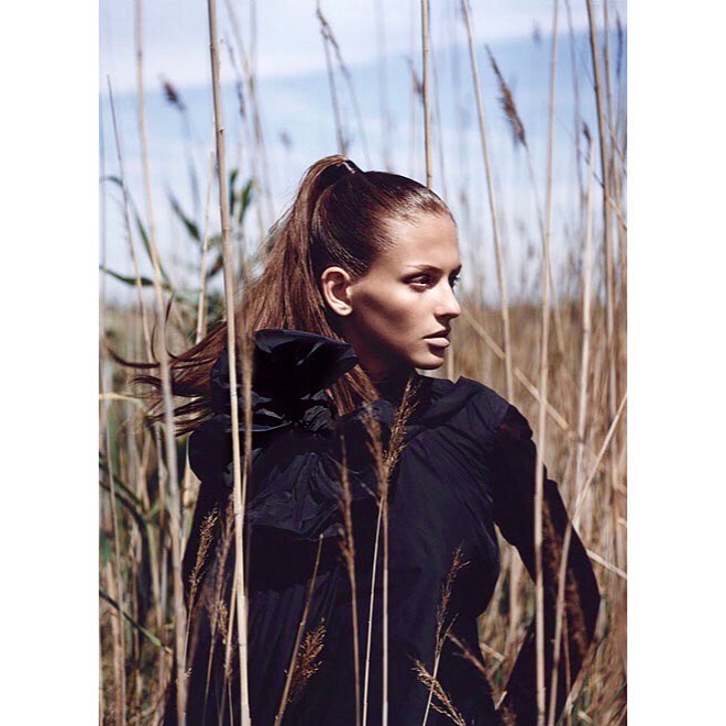 Portrait of a brown-haired woman in a black top standing in a field of wheat-colored tall grass on a sunny day, looking away from the camera.