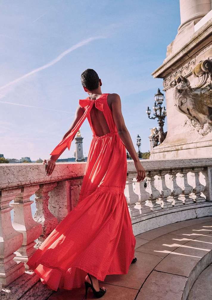 Woman in a red open-back gown with a bow, standing on a stone bridge in Europe with decorative pillars and blue sky. Fashion shot.
