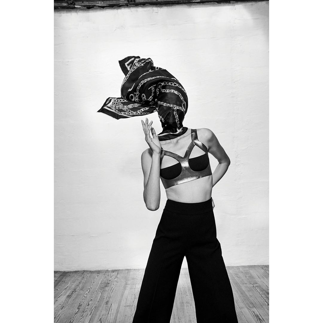Fashionable woman in bra top and wide-leg pants with a patterned scarf draped over her head, posing in a studio with a concrete floor.