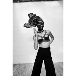 Model wearing a patterned headscarf, strappy cropped top, and black trousers against a textured white wall.