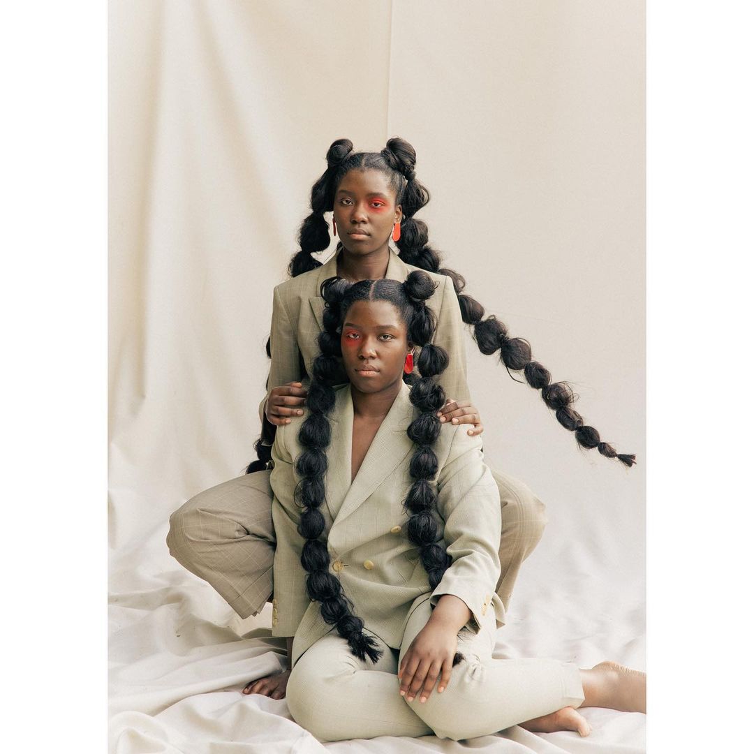 Two Black women pose in matching taupe suits against a pale backdrop, hair styled in long bubble braids, adorned with red makeup and statement earrings.