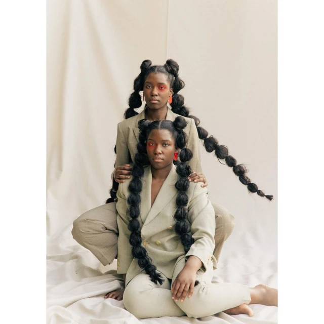 Two Black women pose in matching taupe suits against a pale backdrop, hair styled in long bubble braids, adorned with red makeup and statement earrings.