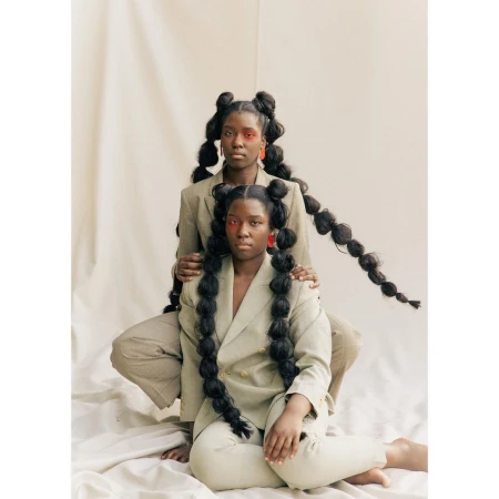 Two Black women pose in matching taupe suits against a pale backdrop, hair styled in long bubble braids, adorned with red makeup and statement earrings.