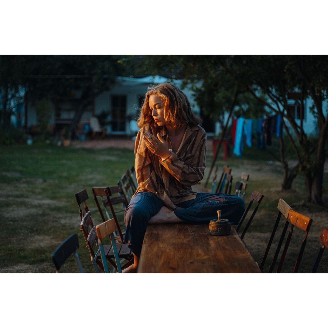 Young woman with curly red hair sits cross-legged on a wooden table in an outdoor garden setting at dusk, holding her hands together.