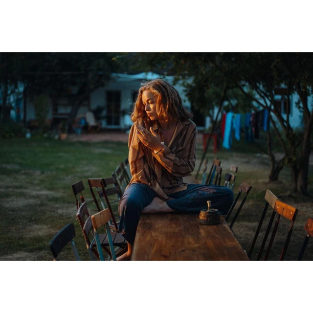 Young woman with curly red hair sits cross-legged on a wooden table in an outdoor garden setting at dusk, holding her hands together.