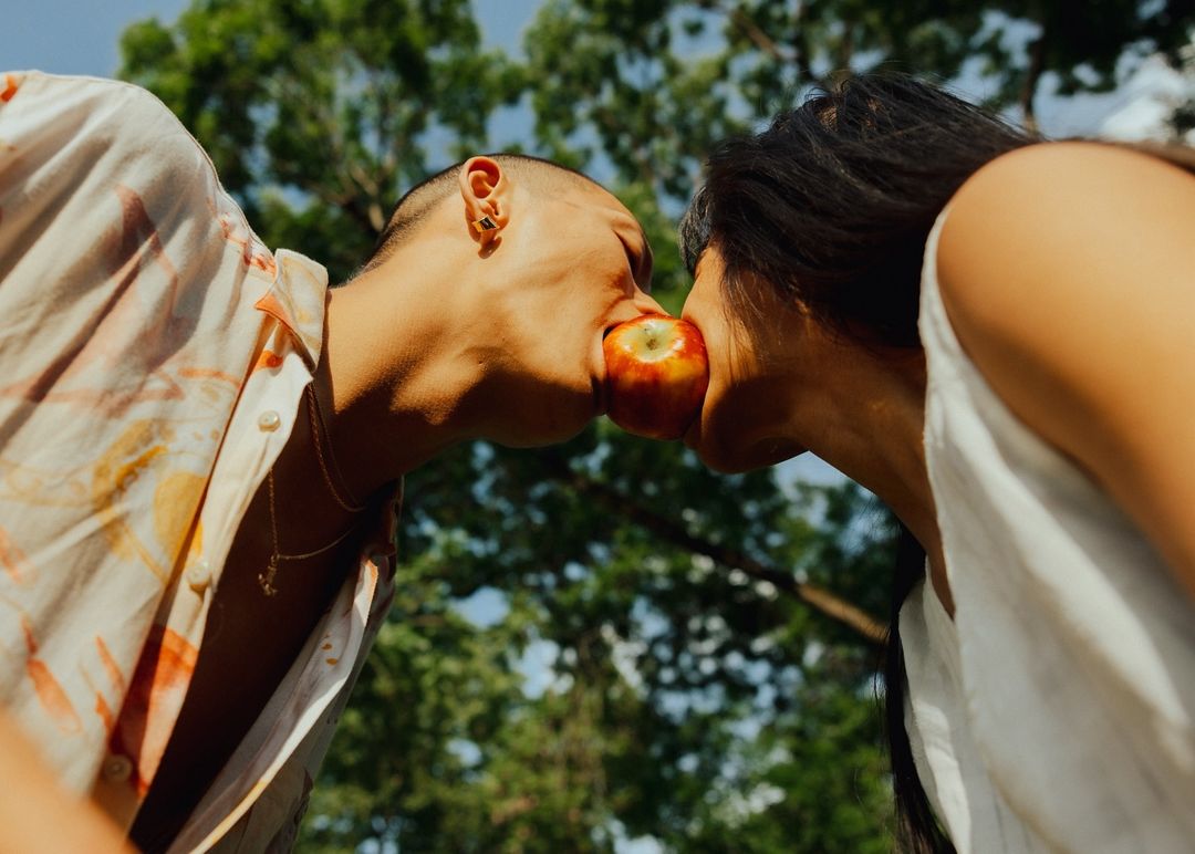 Close-up of two people sharing an apple in an outdoor setting, featuring a couple biting into the same fruit with a leafy background.