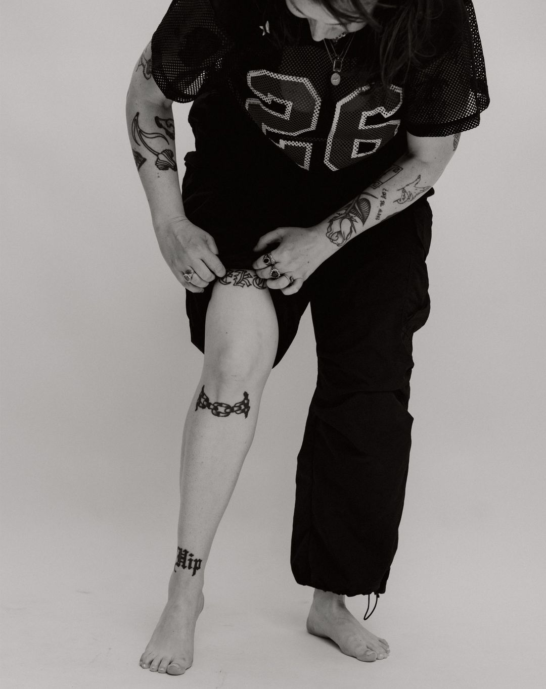 Monochrome shot of an adult wearing a jersey and track pants, showing tattooed leg, and bare feet against a plain background.