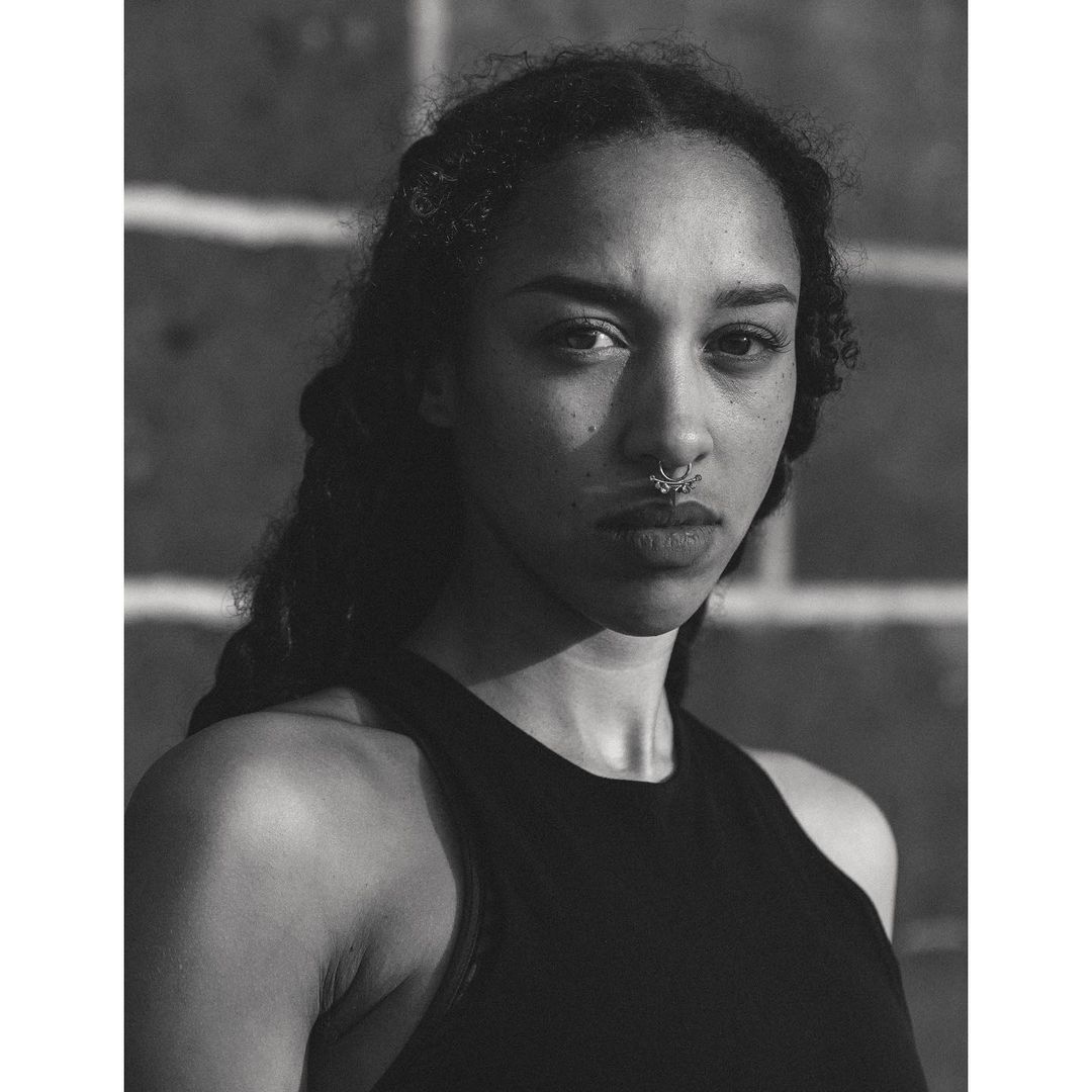 Monochrome portrait of a young woman with dark curly hair and a nose ring, wearing a black tank top against a brick wall background.