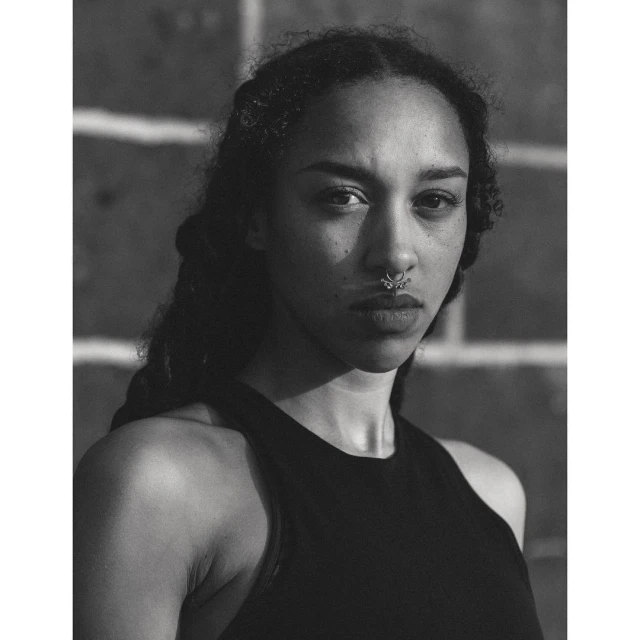 Monochrome portrait of a young woman with dark curly hair and a nose ring, wearing a black tank top against a brick wall background.