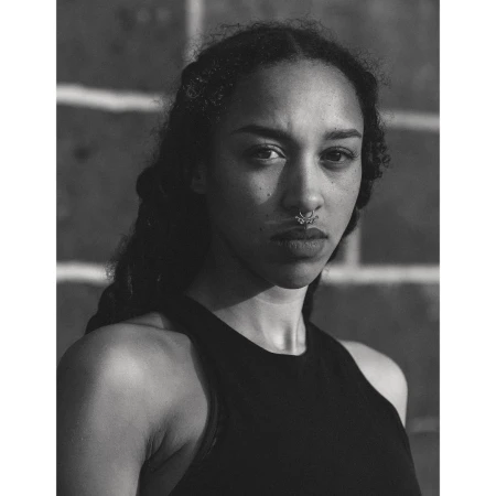 Monochrome portrait of a young woman with dark curly hair and a nose ring, wearing a black tank top against a brick wall background.