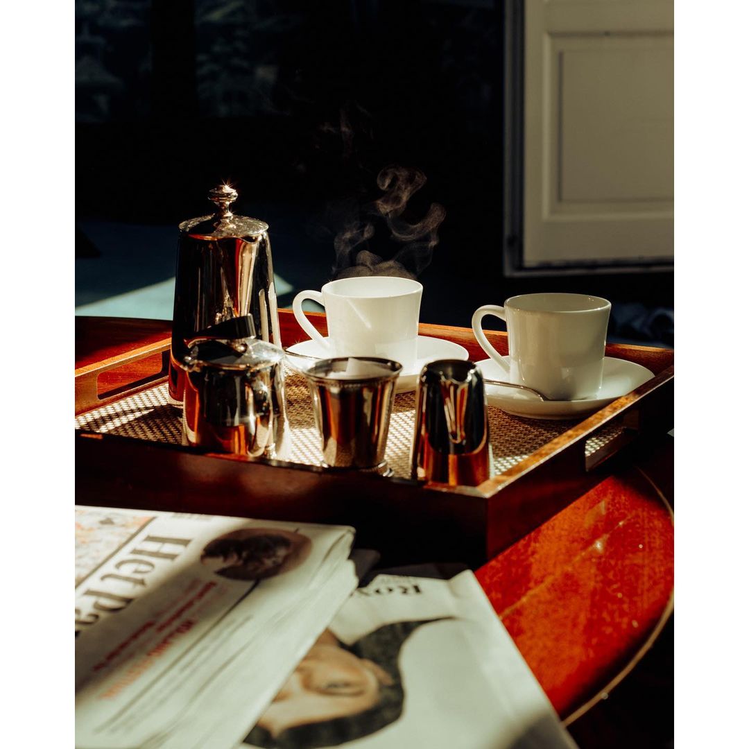 Still life of coffee set with a silver coffee pot, cups, and newspaper on a wooden tray, creating a warm, inviting scene.