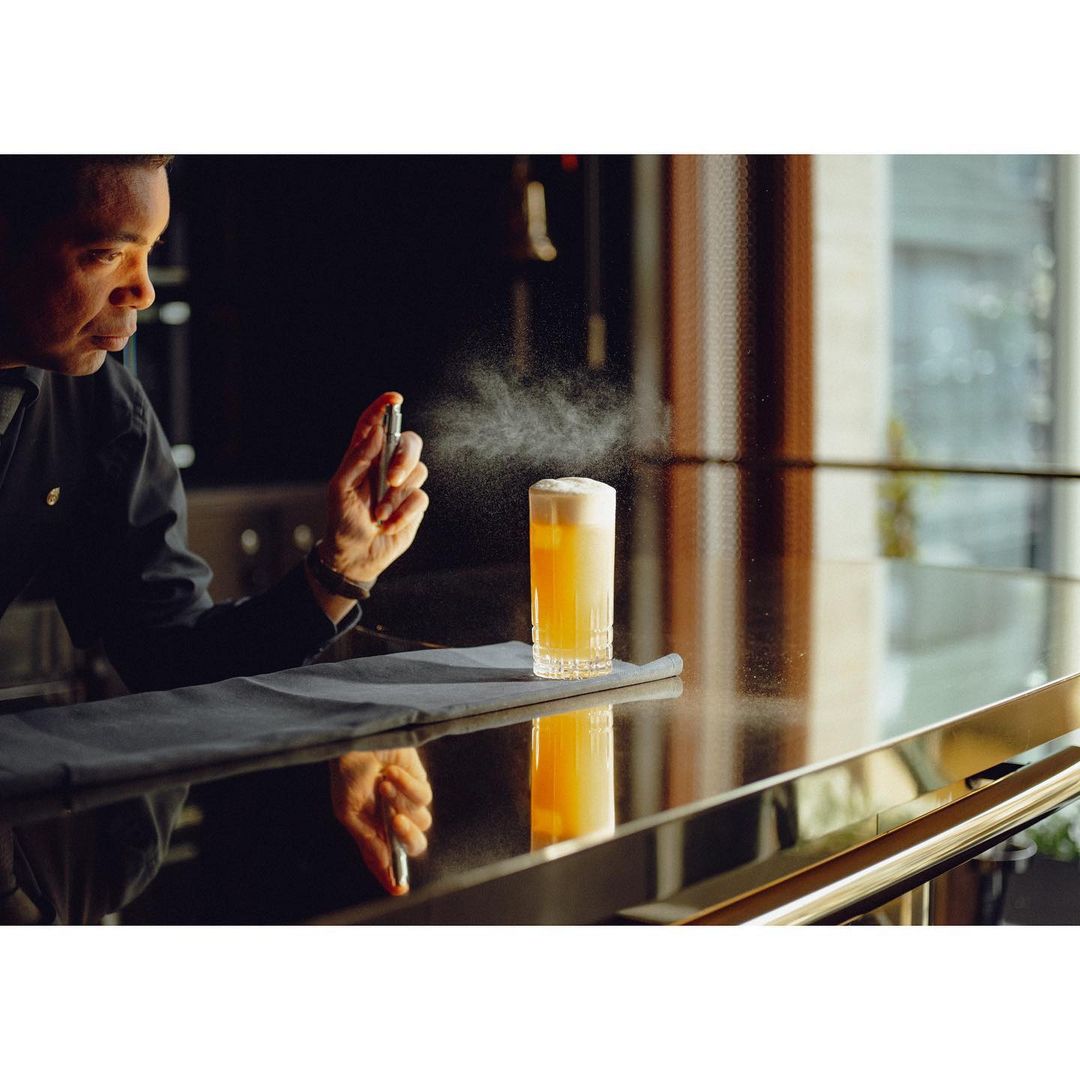 Bartender sprays aromatic mist onto a foamy cocktail on a bar with a reflective surface in warm, subdued lighting. Beverage service.