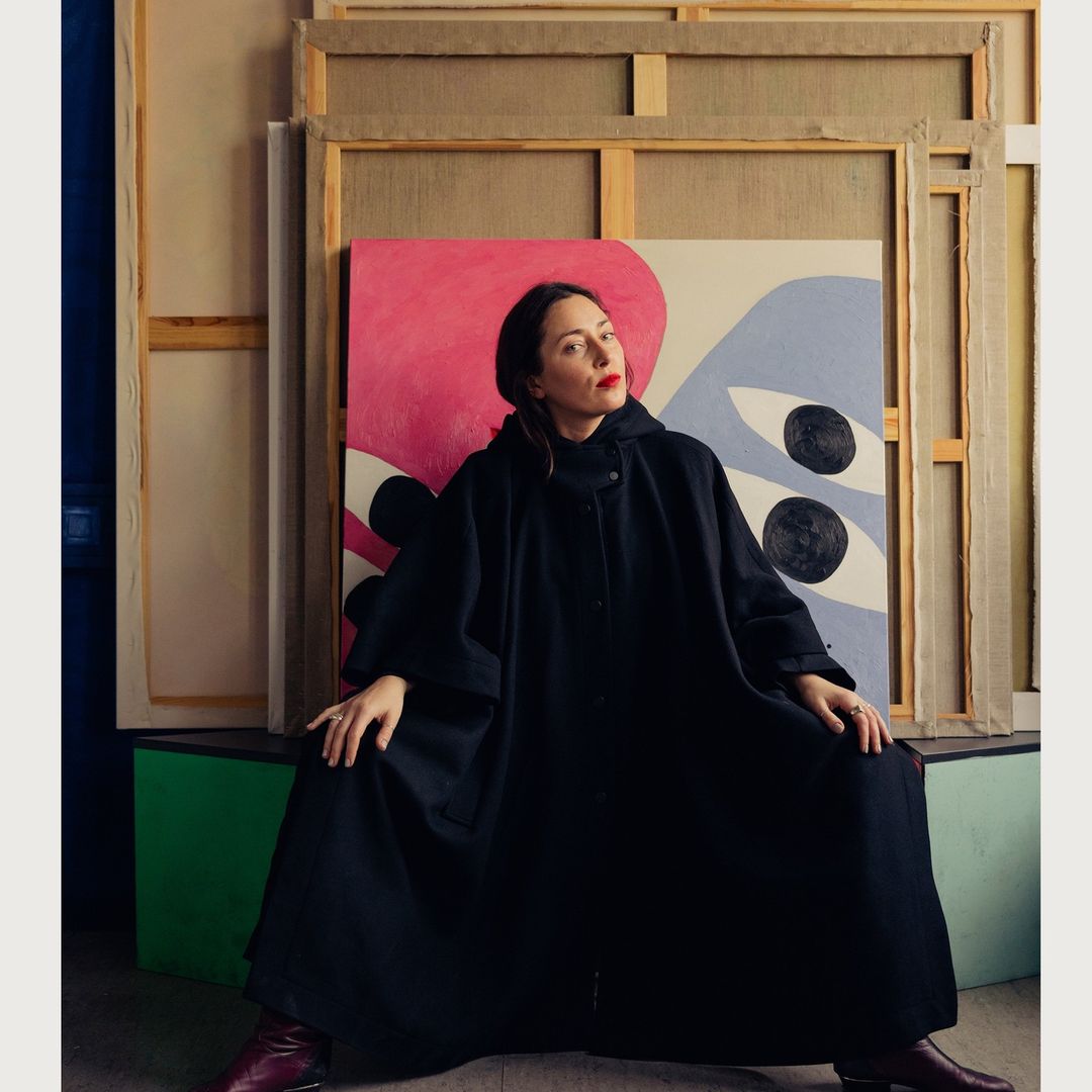 An adult female artist in a black cloak poses in an art studio surrounded by colorful abstract paintings, wearing dark red lipstick.