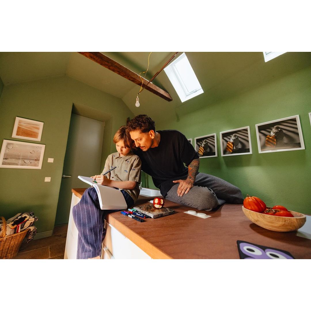 Adult man and child sit on a kitchen island, drawing together. Indoor shot in a room with sage green walls, artwork, and wooden accents.
