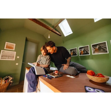 Adult man and child sit on a kitchen island, drawing together. Indoor shot in a room with sage green walls, artwork, and wooden accents.