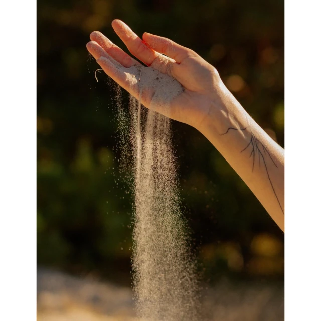 Closeup of an arm and hand as sand pours from fingers, against blurred foliage backdrop; emphasizing textures, light, and natural elements.