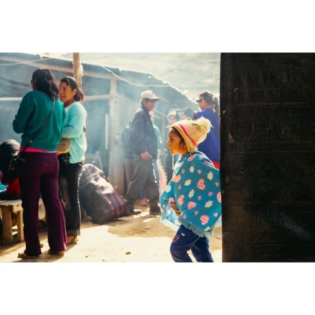 Outdoor market scene featuring a child in a patterned poncho and hat, with a group of adults in the background near a rustic building.