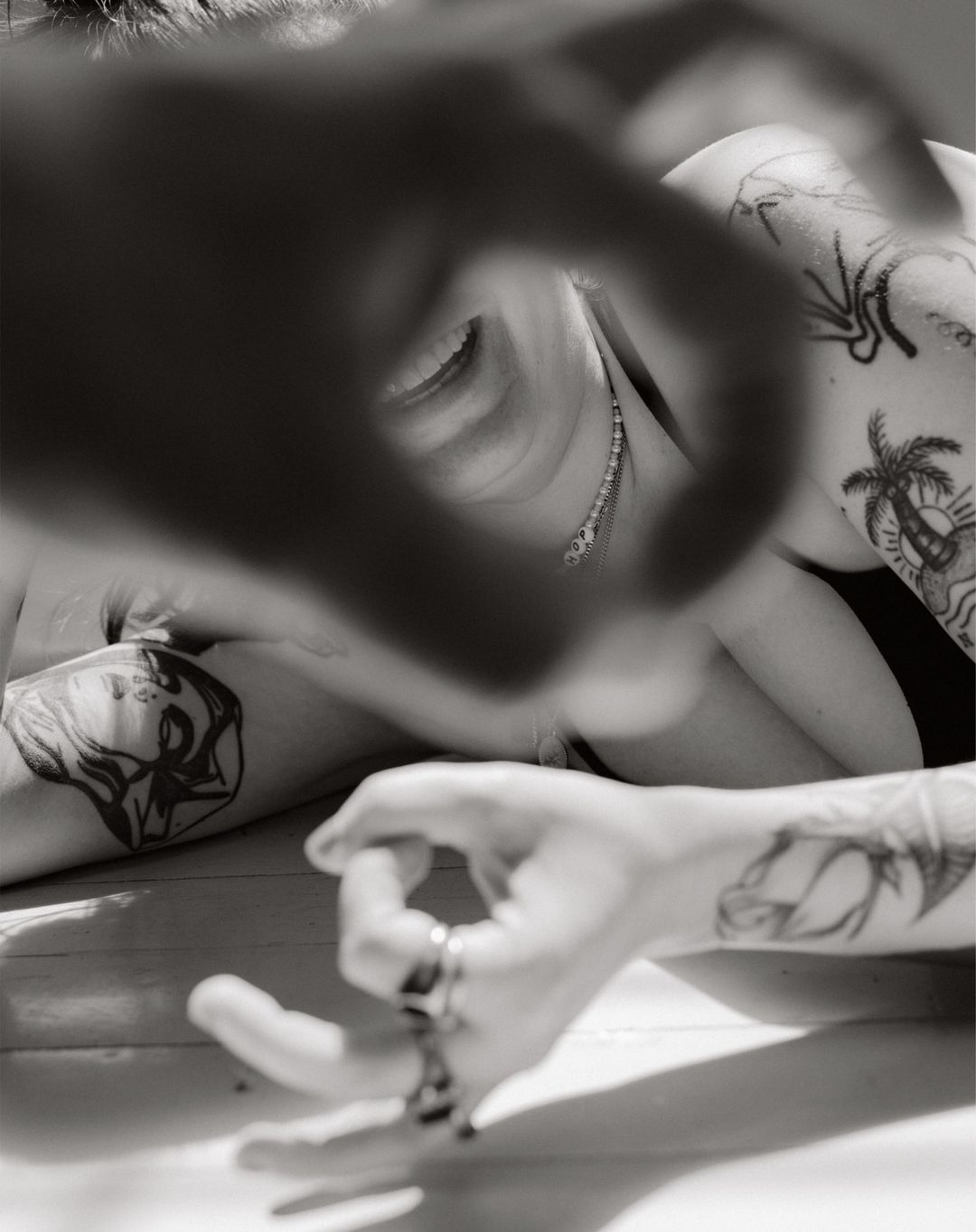 Monochrome close-up of a woman with tattoos making an OK sign around her eye, lying down on a wooden surface in a bright setting.