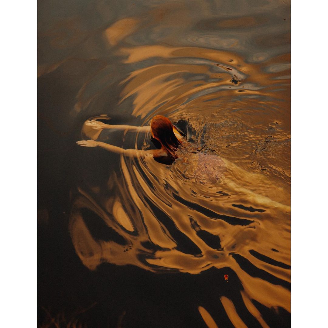 Woman with red hair, back to camera, wading through dark water with rippled surface, warm colour palette, slight motion blur.