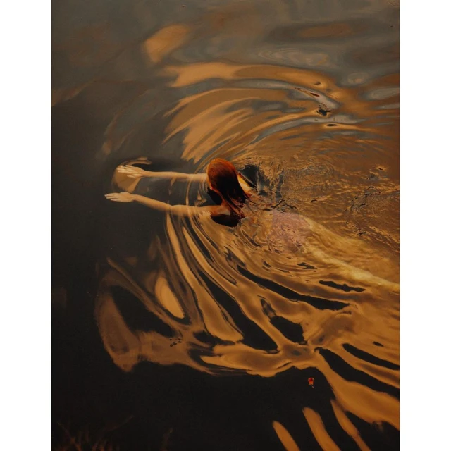 Woman with red hair, back to camera, wading through dark water with rippled surface, warm colour palette, slight motion blur.