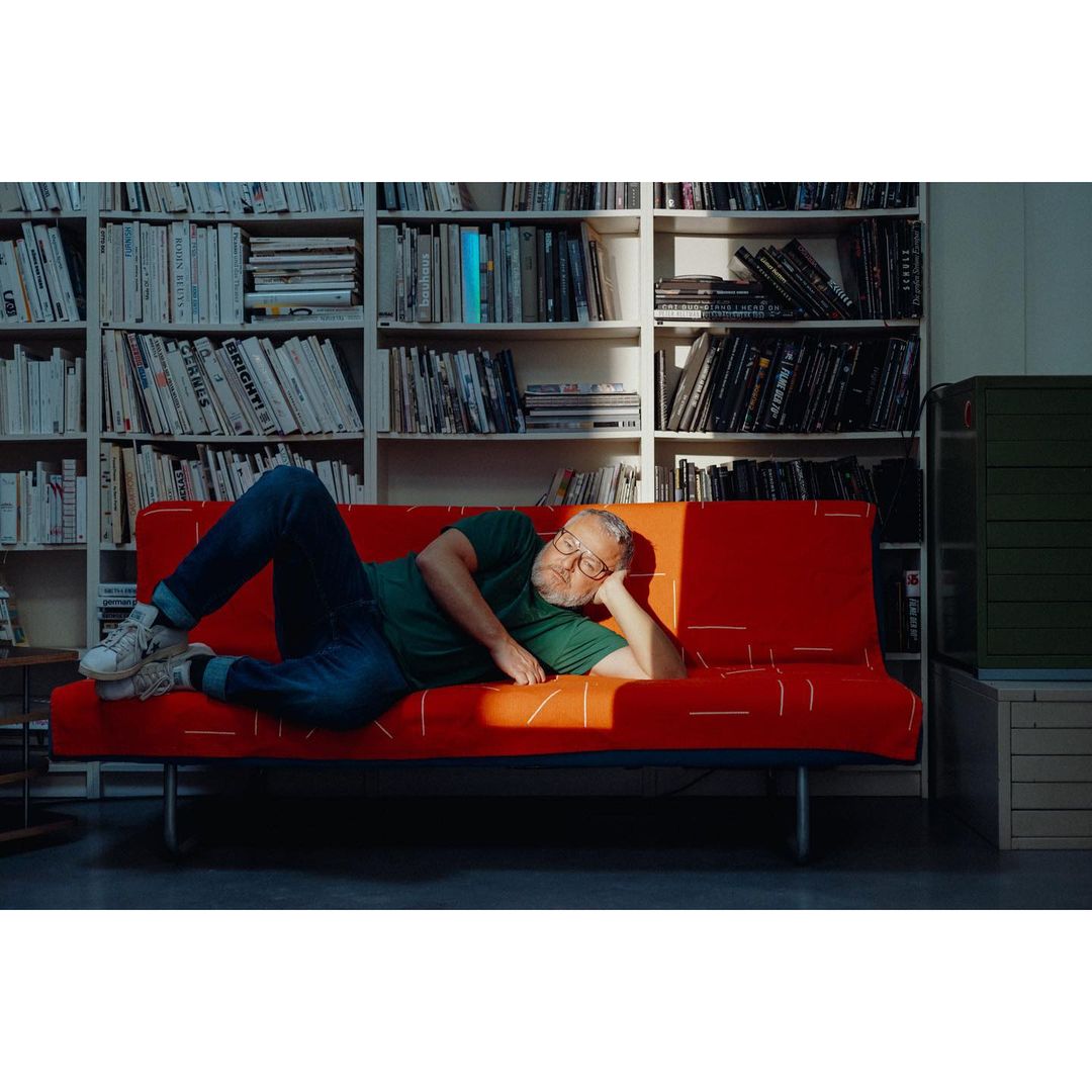 Man relaxes on a red patterned couch in a room filled with books. The setting gives a cozy, indoor, and artistic vibe.