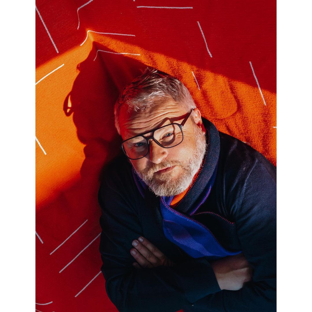 Portrait of a man with glasses in dark clothing against a vibrant orange patterned backdrop, showcasing a modern and unique aesthetic.