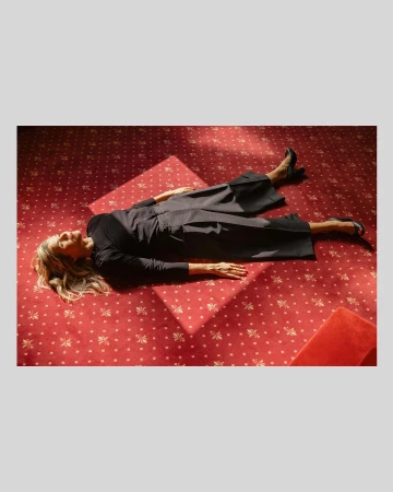 Woman lying flat on a red patterned carpet wearing a dark top and dark trousers captured by Ramon Haindl