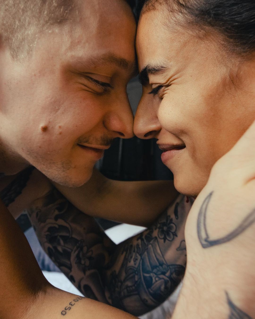 Close-up shot of a smiling man and woman, facing each other with eyes closed and foreheads touching, adorned with tattoos on their arms.