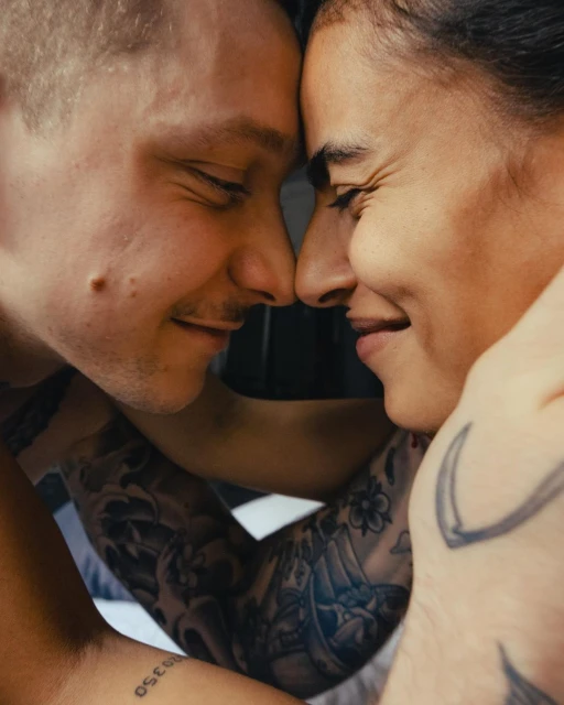 Close-up shot of a smiling man and woman, facing each other with eyes closed and foreheads touching, adorned with tattoos on their arms.