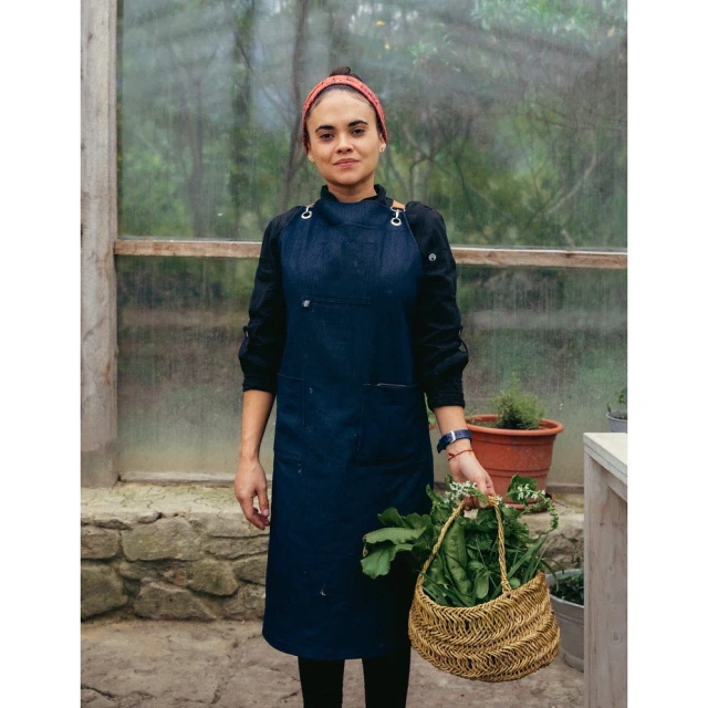 A woman in a denim apron stands in a greenhouse holding a basket of fresh vegetables, bathed in soft light. 
