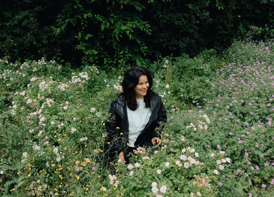 A woman with dark hair in a white shirt and black jacket smiles while standing in a field of mixed pink, white, and yellow flowers.
