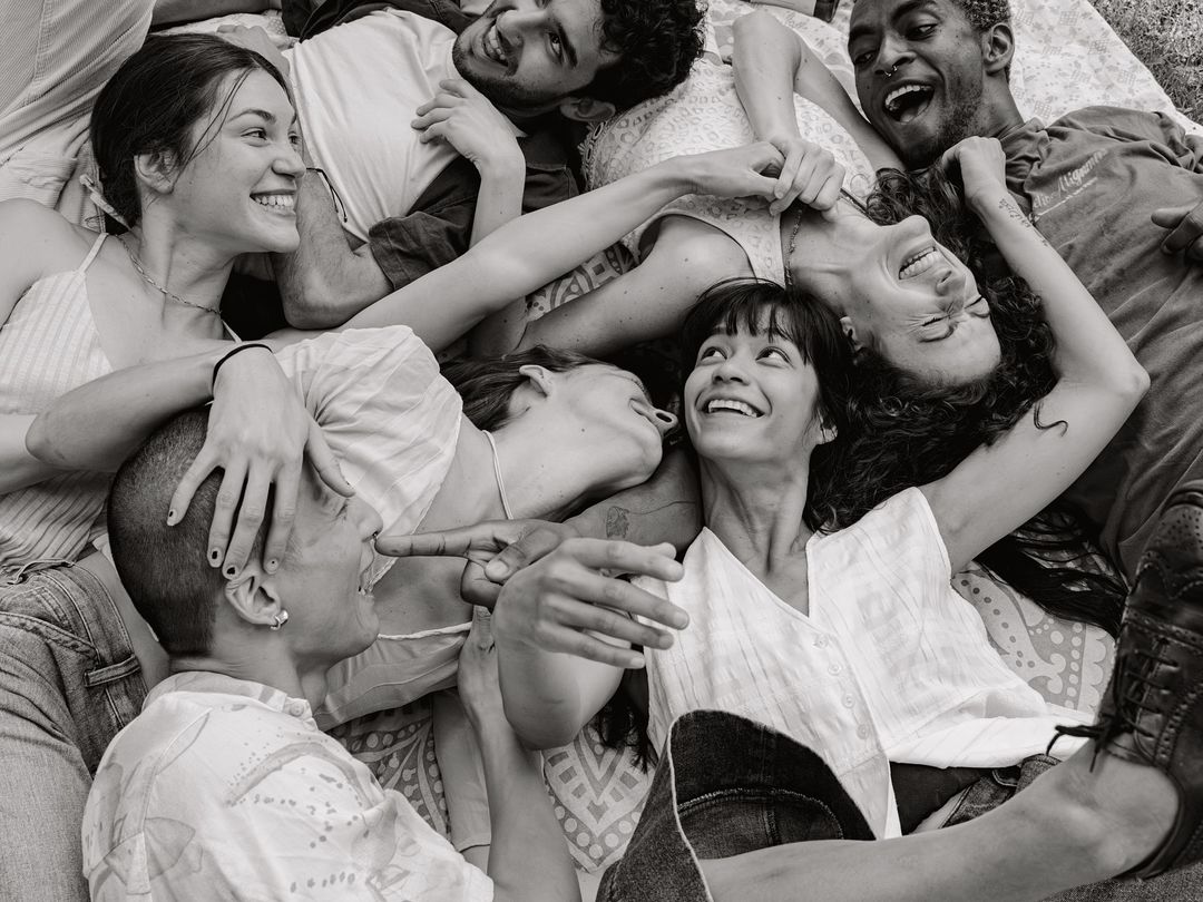 Black and white, high-angle view of a laughing diverse group of adults lying in a pile on a patterned blanket outdoors.