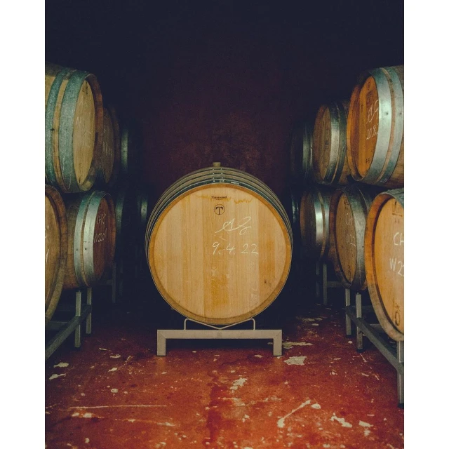 Image of a wine barrel in a cellar with handwriting on the front, surrounded by other wooden barrels in the background.