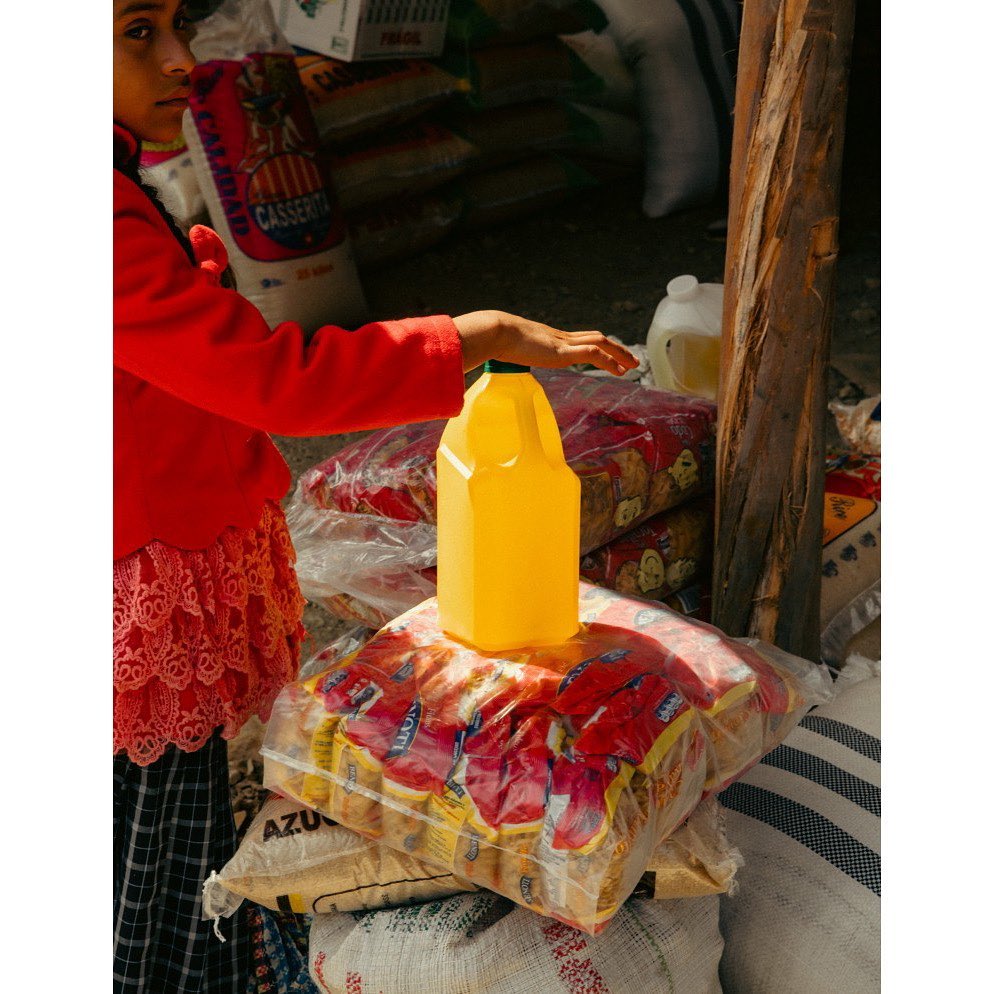 Young person in red stands behind a stack of packaged goods, with a yellow juice container on top, in an outdoor market setting.